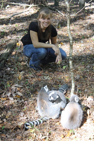 Katie Grogan in a black t-shirt and jeans crouching in the forest next to three ring-tailed lemurs